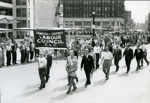 Delegation from the Hamilton & District Labour Council marching west at Market Square. Mayor Victor Kennedy Copps is in the white hat marching behind the banner. King William Street and the Lister building are in the background. Delegation from the Hamilton & District Labour Council marching west at Market Square. Mayor Victor Kennedy Copps is in the white hat marching behind the banner. King William Street and the Lister building are in the background.